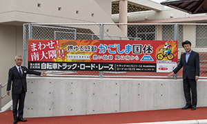 役場敷地内に設置された看板と第一生命保険株式会社鹿屋営業オフィス部長寺林宏泰さん（写真右）